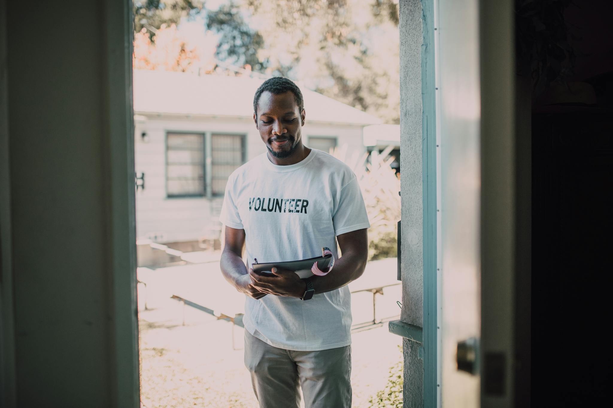 A man wearing a volunteer t-shirt stands by a doorway holding a tablet, engaged in a community event.